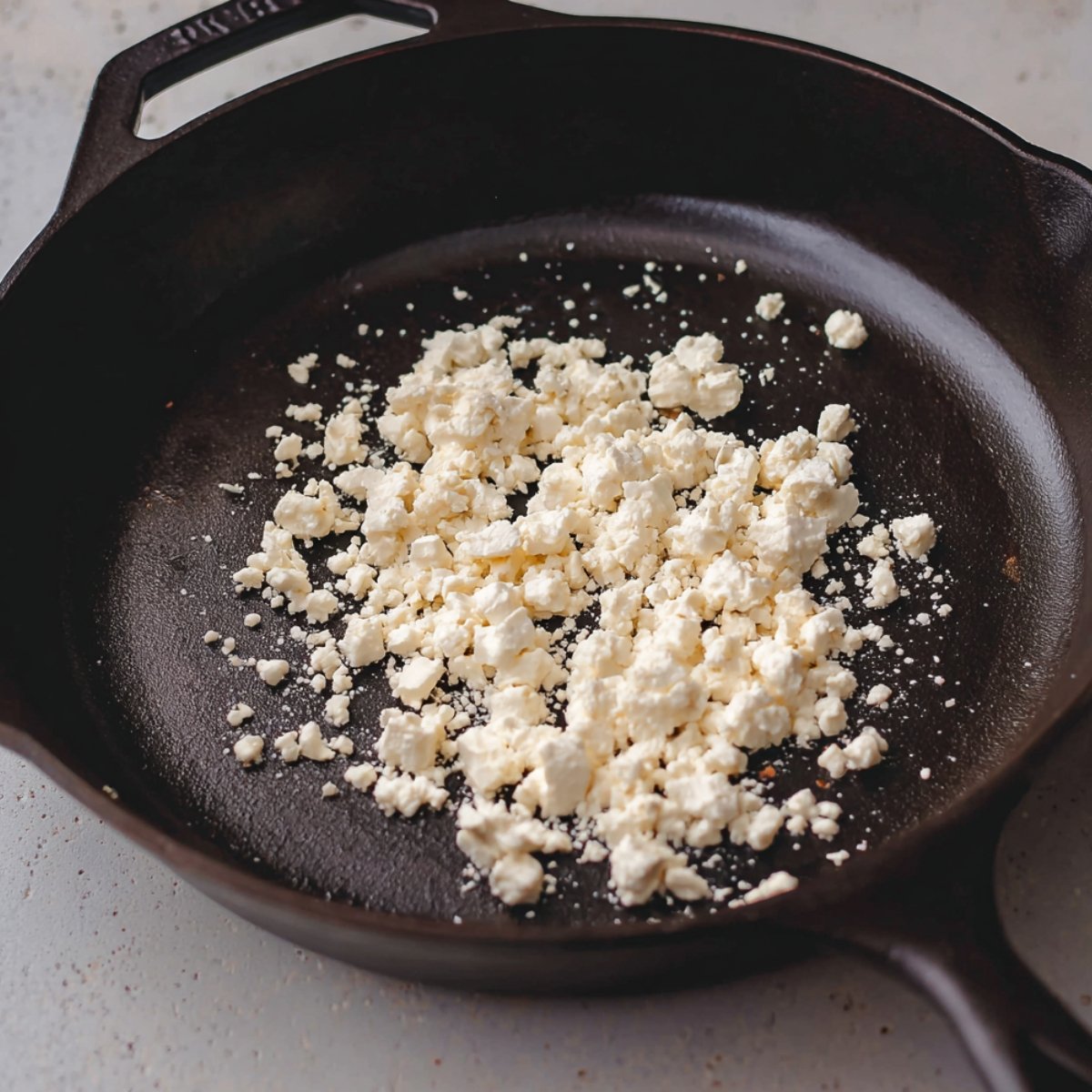 A skillet with crumbled feta cheese cooking in it, showing a textured and slightly browned appearance.