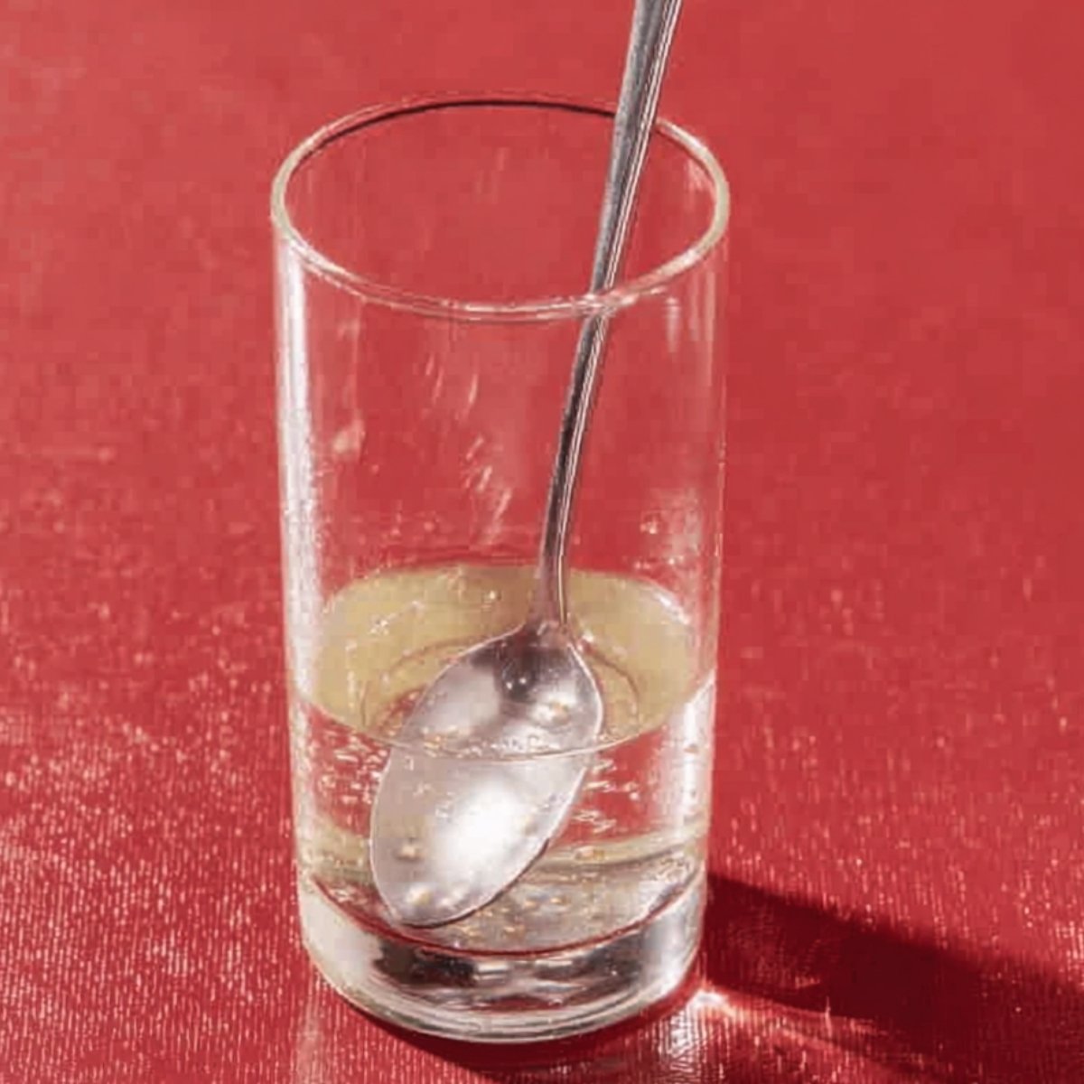 A glass with a spoon resting inside, filled with a clear liquid and a small amount of dissolved sugar at the bottom. The background is red.