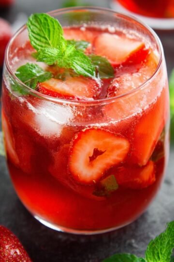 A glass of strawberry iced tea garnished with mint leaves and fresh strawberries on a dark surface.