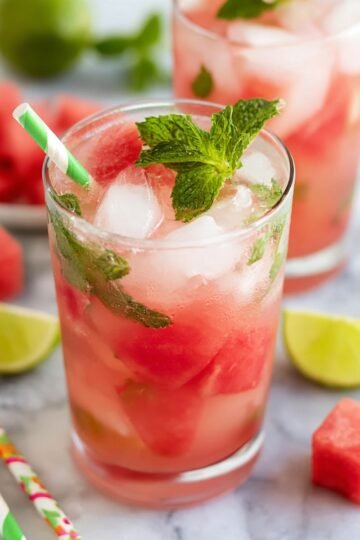A refreshing watermelon mojito served in a glass, garnished with mint leaves and ice cubes. Straws in festive colors are placed nearby, and the drink is surrounded by watermelon cubes and lime wedges.