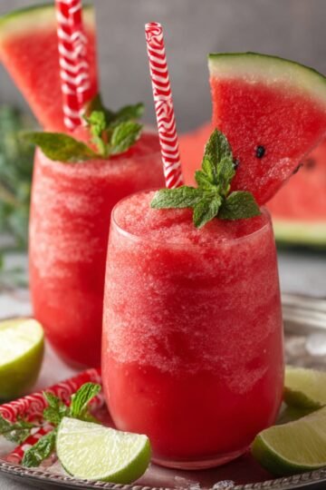 Two glasses of refreshing watermelon slushie garnished with mint leaves, a slice of watermelon, and striped straws, served on a silver tray with lime wedges and fresh mint.